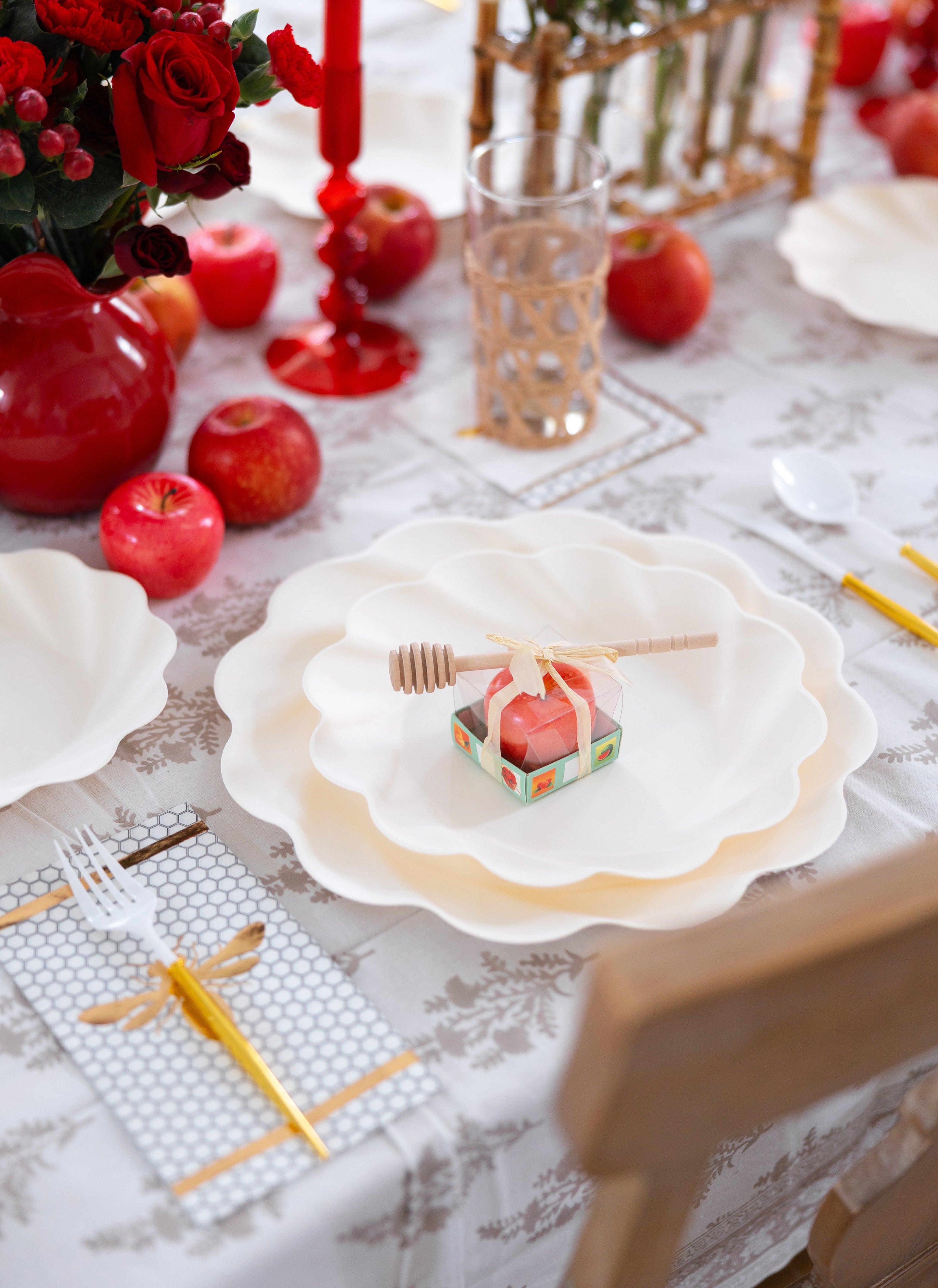 A decorative table setting featuring the Sophistiplate LLC Rosh Tablescape for 16 Guests: Eco Cream Honeybee, with a honey dipper on a wrapped apple, red apples, red flowers, red candles, gold cutlery, and a patterned tablecloth.