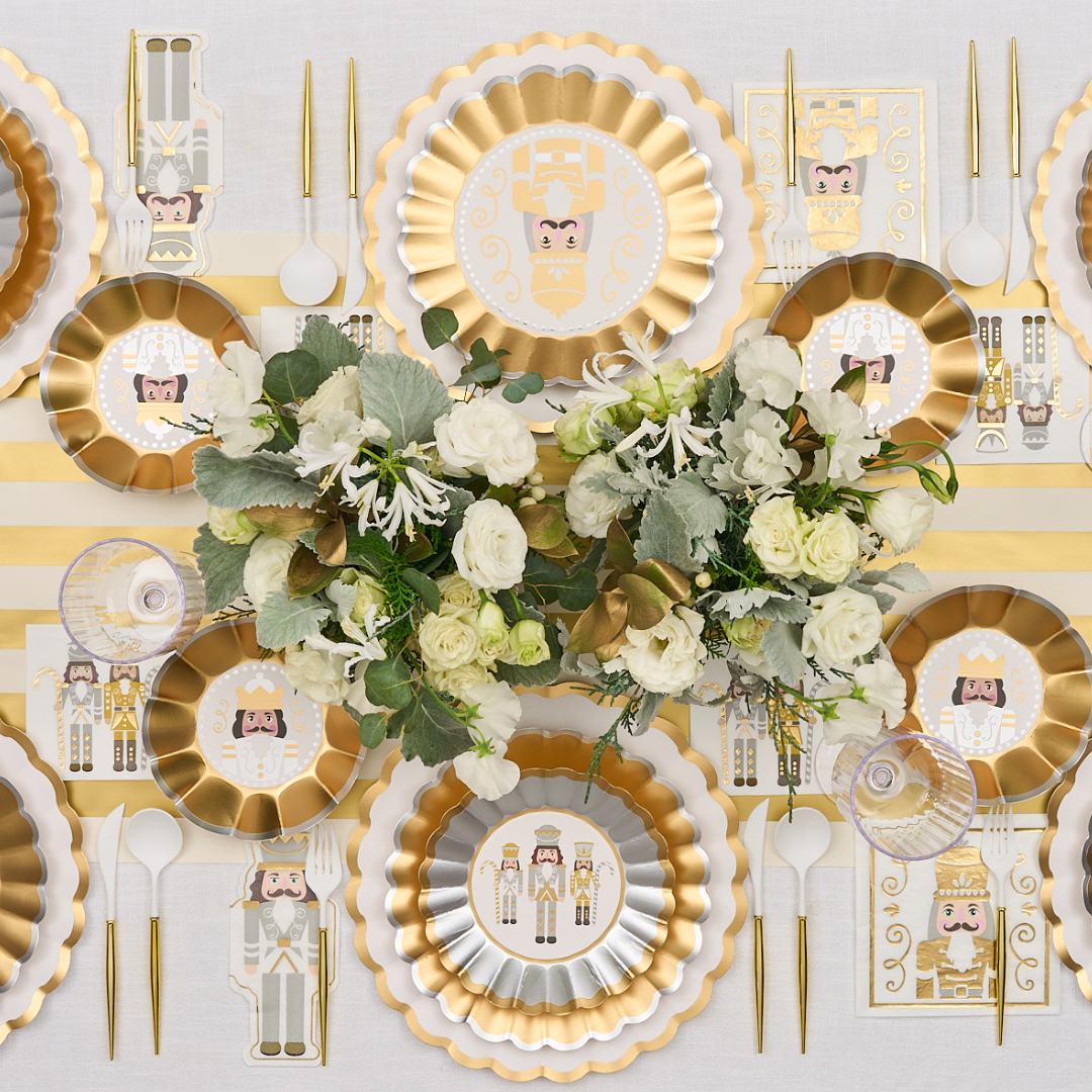 The Gilded Nutcracker Table Setting by Sophistiplate features festive plates, gold cutlery, striped napkins, and a white-green floral centerpiece—an elegant holiday-inspired disposable tableware set for effortless entertaining.