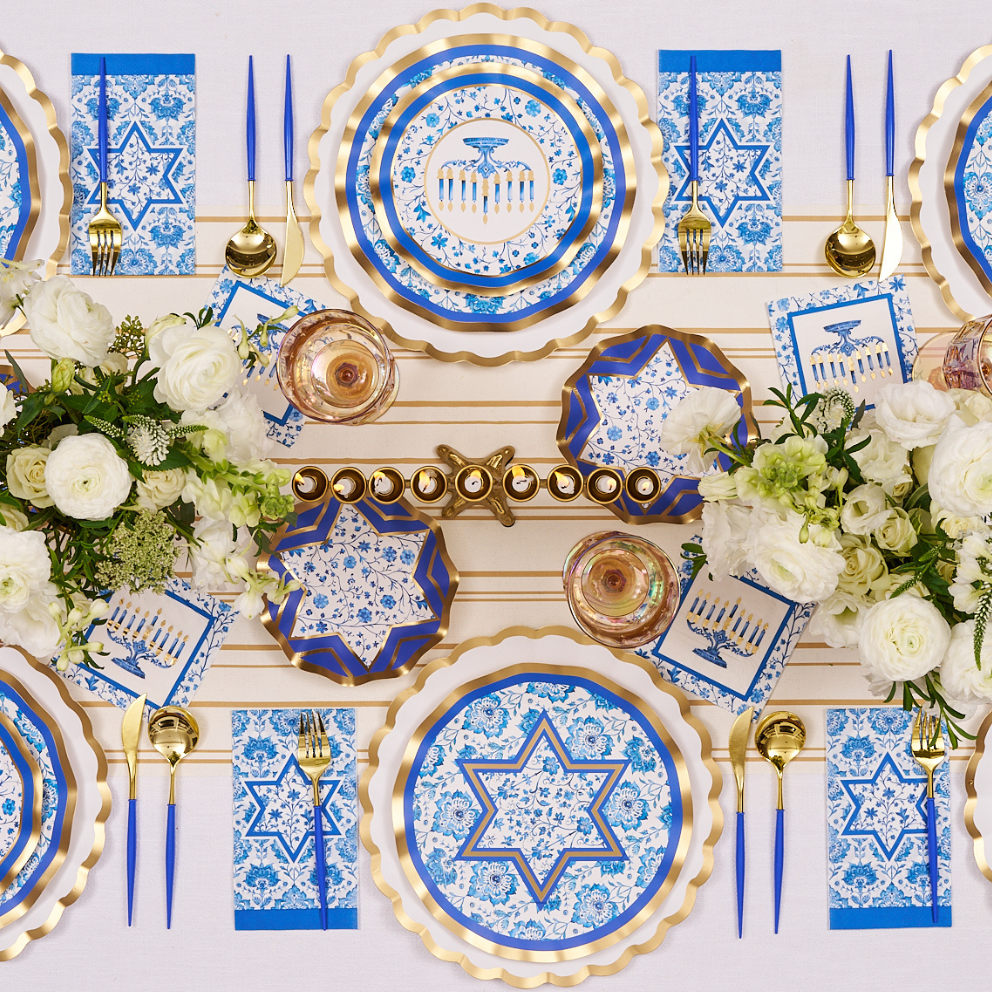 Create a stunning Hanukkah celebration with the Hanukkah Toile Table Setting from Sophistiplate, featuring blue and gold designer paper plates adorned with Stars of David and menorah motifs, elegant gold cutlery, white floral accents, and a menorah centerpiece with candles—all beautifully arranged on a crisp white tablecloth for a stylish and festive tablescape.