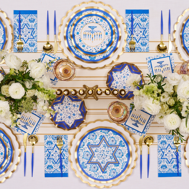 Create a stunning Hanukkah celebration with the Hanukkah Toile Table Setting from Sophistiplate, featuring blue and gold designer paper plates adorned with Stars of David and menorah motifs, elegant gold cutlery, white floral accents, and a menorah centerpiece with candles—all beautifully arranged on a crisp white tablecloth for a stylish and festive tablescape.