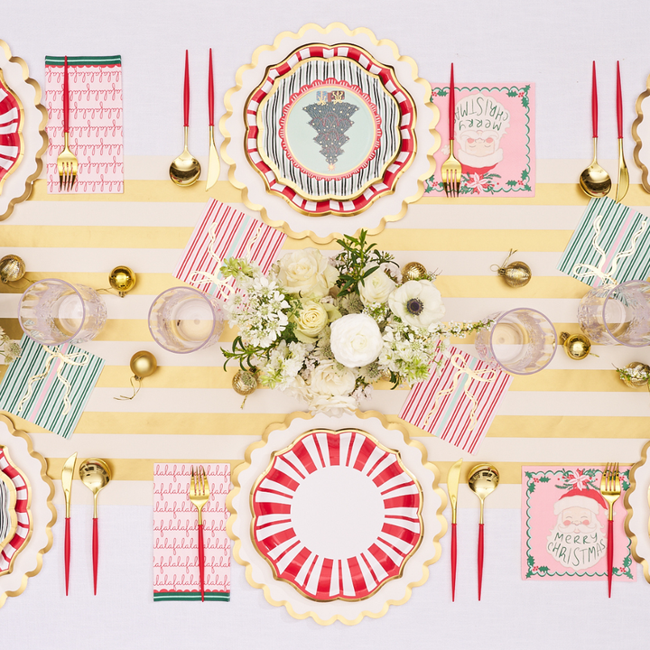 A festive table setting viewed from above features red, gold, and white decor, Sophistiplate’s Tis the Season Dinner Plates (8pk) with a striped Christmas design, gold cutlery, holiday-themed napkins, and a floral centerpiece.