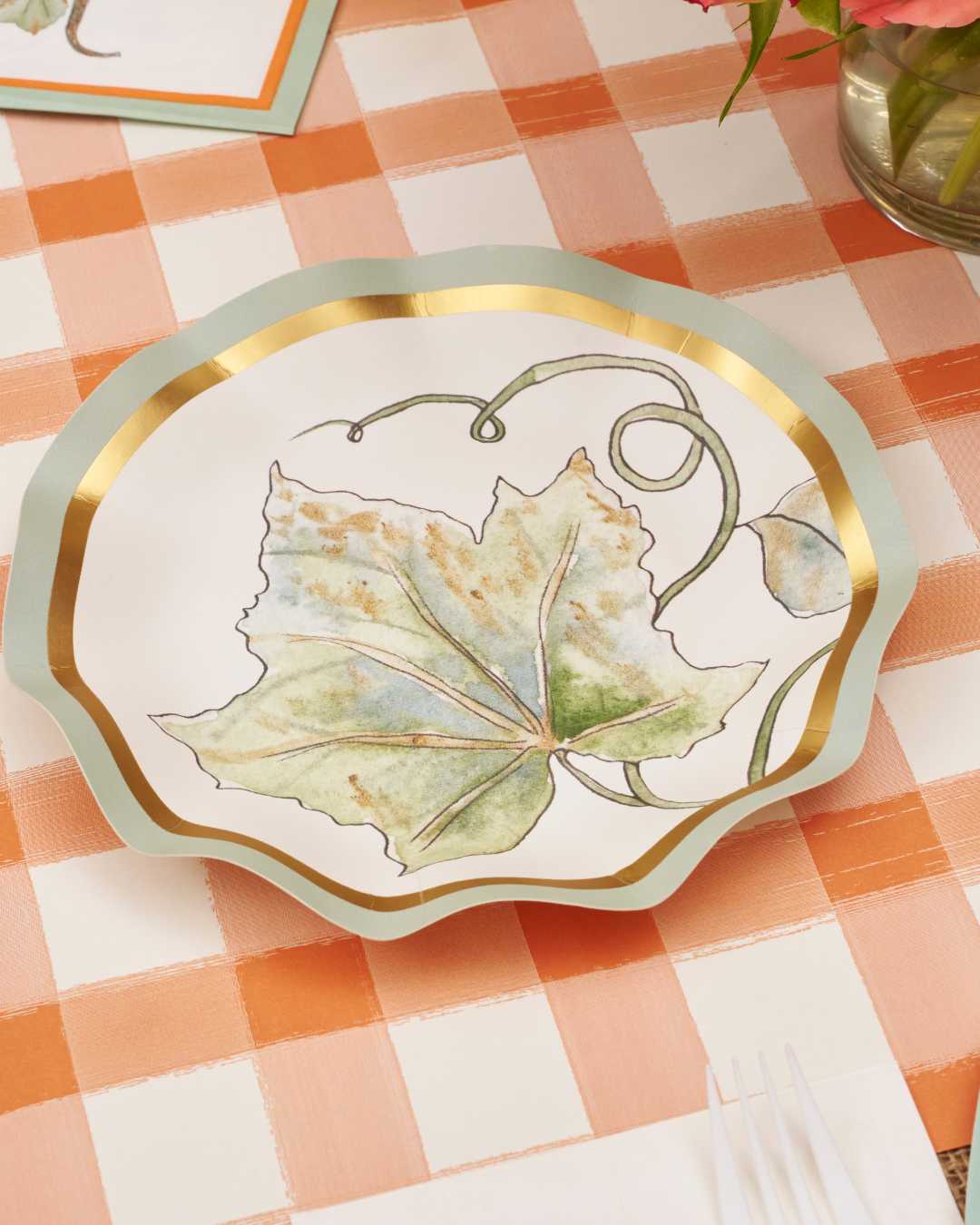 The Sophistiplate Bright & Thankful Table Setting features a decorative plate with a scalloped edge and gold rim, adorned with watercolor leaves and vines. Perfect for autumn, it’s shown on an orange and white checkered tablecloth.