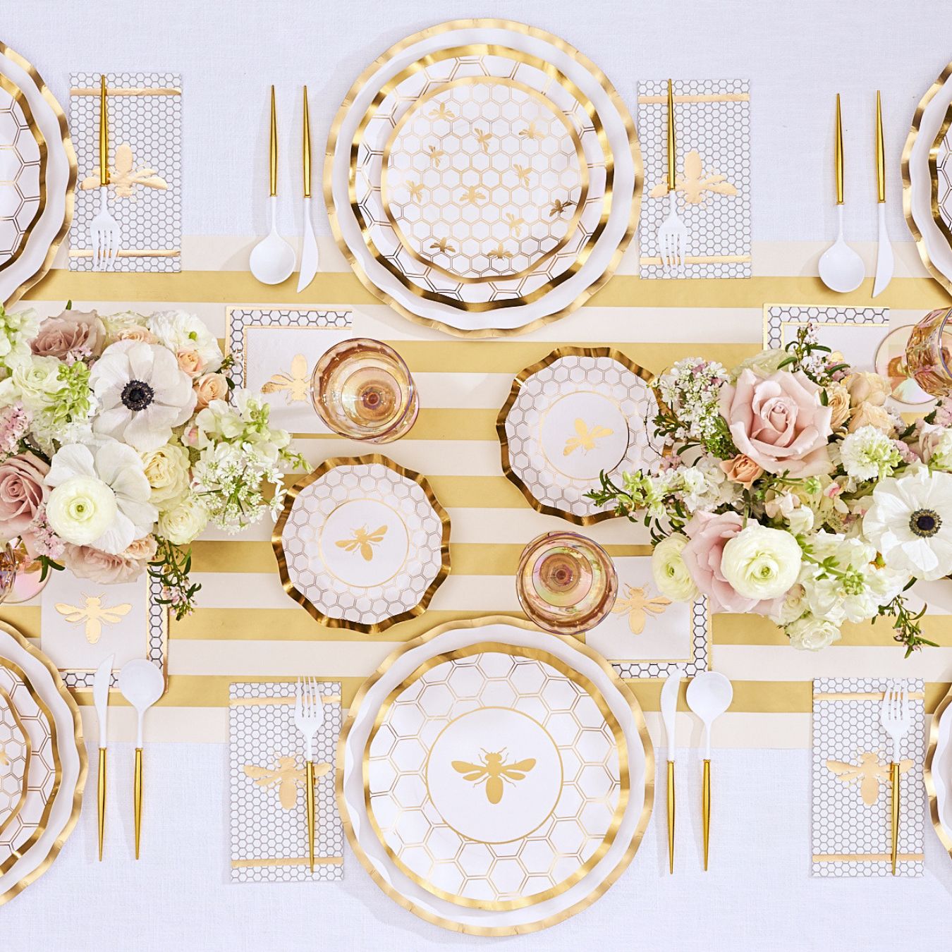 The Sophistiplate White Honeybee Table Setting features gold and white bee-themed plates, gold-rimmed paper plates, elegant gold utensils, a striped table runner, floral centerpieces with roses and ranunculus, and honeycomb-patterned napkins to create an elegant party tableware display.