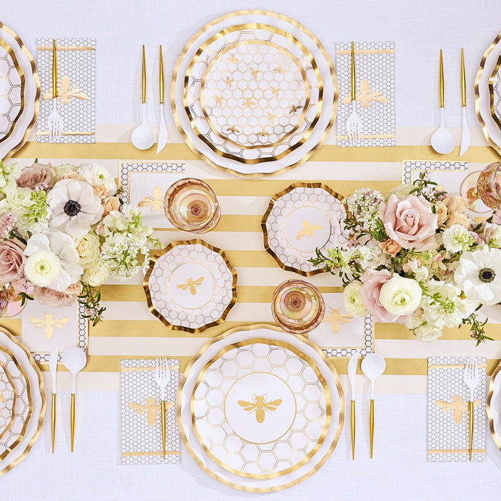 The Sophistiplate White Honeybee Table Setting features gold and white bee-themed plates, gold-rimmed paper plates, elegant gold utensils, a striped table runner, floral centerpieces with roses and ranunculus, and honeycomb-patterned napkins to create an elegant party tableware display.