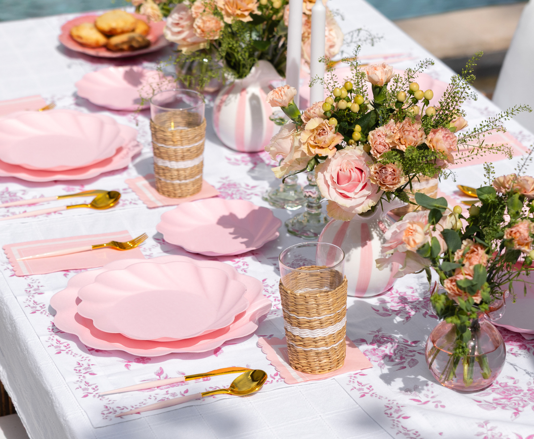 The Eco-Friendly Table Setting- Blush by Sophistiplate LLC features pink scalloped plates, gold cutlery, woven glasses, floral napkins, and vases of pink roses for an elegant, festive outdoor celebration.
