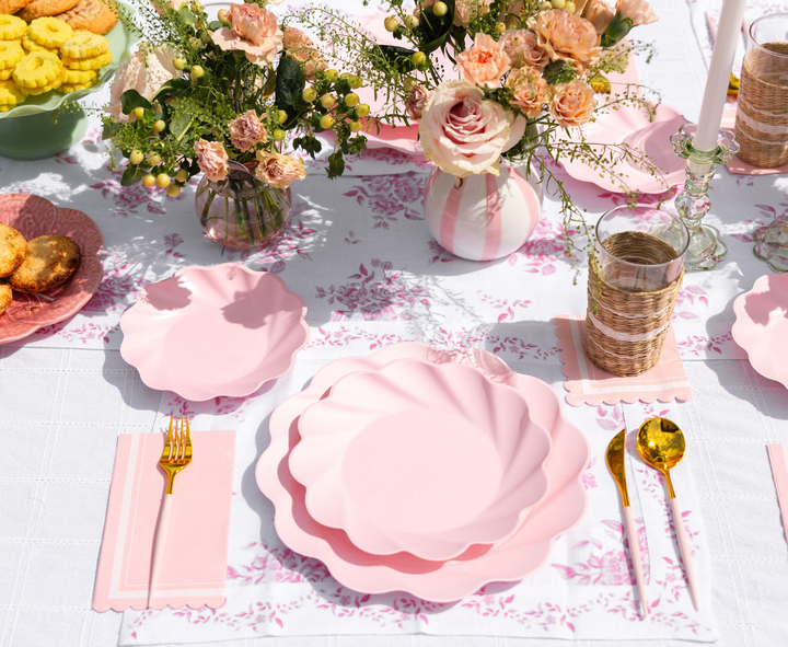 The Sophistiplate LLC Eco-Friendly Table Setting- Blush features disposable plates, gold cutlery, pink napkins, floral vases, and glass cups with woven holders on a white and pink floral tablecloth—perfect for a spring meal.