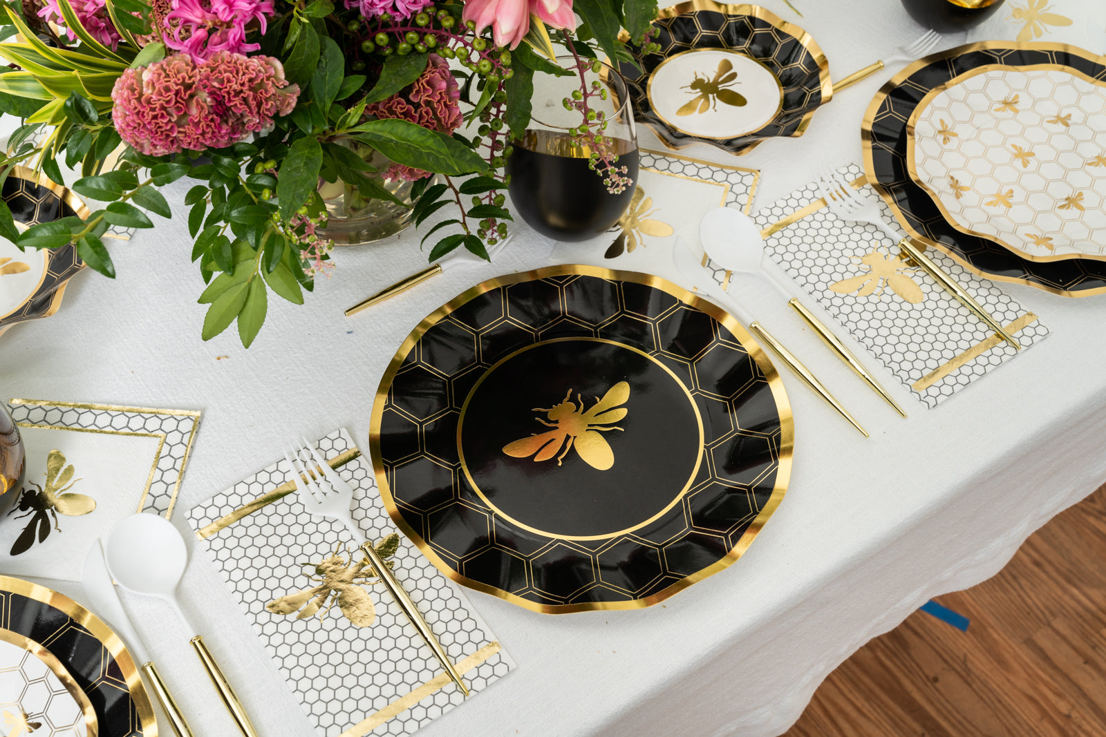 A meal table features Sophistiplate Honeybee Paper Guest Towels, black and gold bee-themed plates, gold cutlery, metallic gold foil napkins, and pink flowers on a white tablecloth.