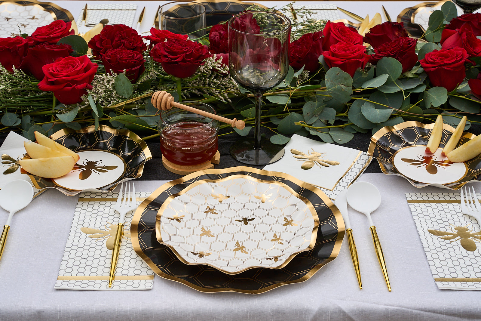 A festive table features Sophistiplate’s Honeybee Paper Cocktail Napkins, honeycomb bee plates, gold cutlery, a jar of honey with dipper, sliced apples, and a centerpiece of red roses and greenery.