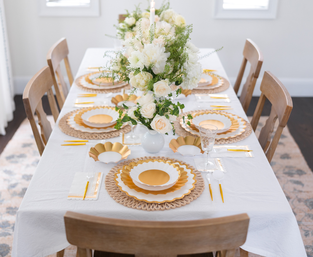 The Gold & White Table Setting by Sophistiplate LLC seats six, featuring designer white and gold disposable plates, gold cutlery, crystal glasses, and a white tablecloth with floral centerpieces in white vases.