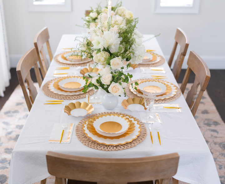 The Gold & White Table Setting by Sophistiplate LLC seats six, featuring designer white and gold disposable plates, gold cutlery, crystal glasses, and a white tablecloth with floral centerpieces in white vases.