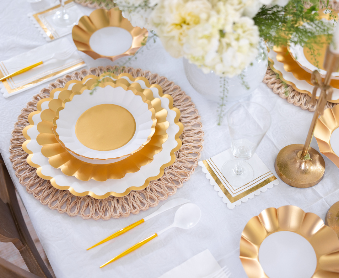 A close-up of the Gold & White Table Setting by Sophistiplate LLC, featuring gold and white scalloped plates, gold flatware, clear glassware, decorative gold-trimmed napkins, a floral centerpiece, and a white tablecloth.