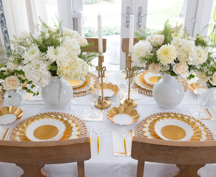 The Gold & White Table Setting by Sophistiplate LLC features designer disposable plates, gold cutlery, white napkins, candles in gold holders, and white flower arrangements for an elegant, festive dining atmosphere.