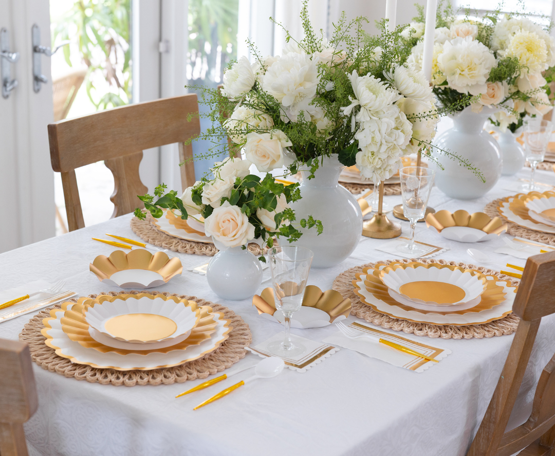 The Gold & White Table Setting by Sophistiplate LLC features designer disposable plates in white and gold, yellow-handled cutlery, glasses, white napkins, floral centerpieces in white vases, and wooden chairs elegantly arranged.