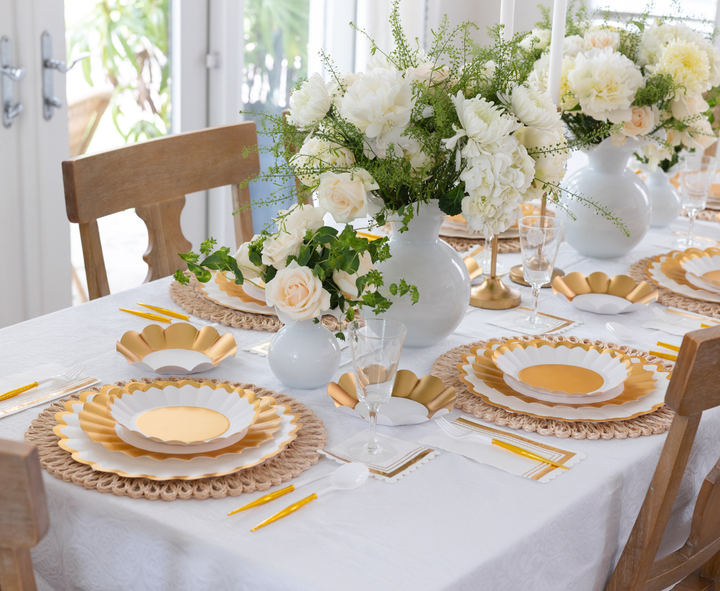 The Gold & White Table Setting by Sophistiplate LLC features designer disposable plates in white and gold, yellow-handled cutlery, glasses, white napkins, floral centerpieces in white vases, and wooden chairs elegantly arranged.