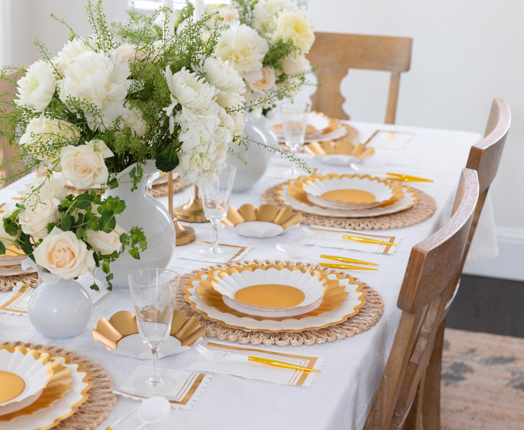 The Gold & White Table Setting by Sophistiplate LLC features designer disposable plates, yellow cutlery, and crystal glasses. A white floral and greenery centerpiece sits on a white cloth, with wooden chairs completing the look.