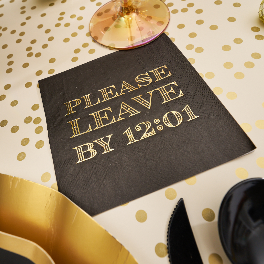 Midnight Sparkle Paper Cocktail Napkin Please Leave By 12:01 /26pk with black and gold-foiled text resting on a gold polka-dot tablecloth.