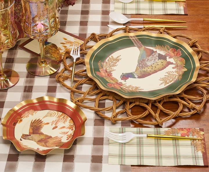 A festive table setting featuring Sophistiplate Woodland Harvest Dinner Plates (8 pack) adorned with pheasants and autumn leaves, paired with plaid napkins, plastic utensils, a woven placemat, and a brown-checkered tablecloth. Two crystal glasses complete the ensemble.