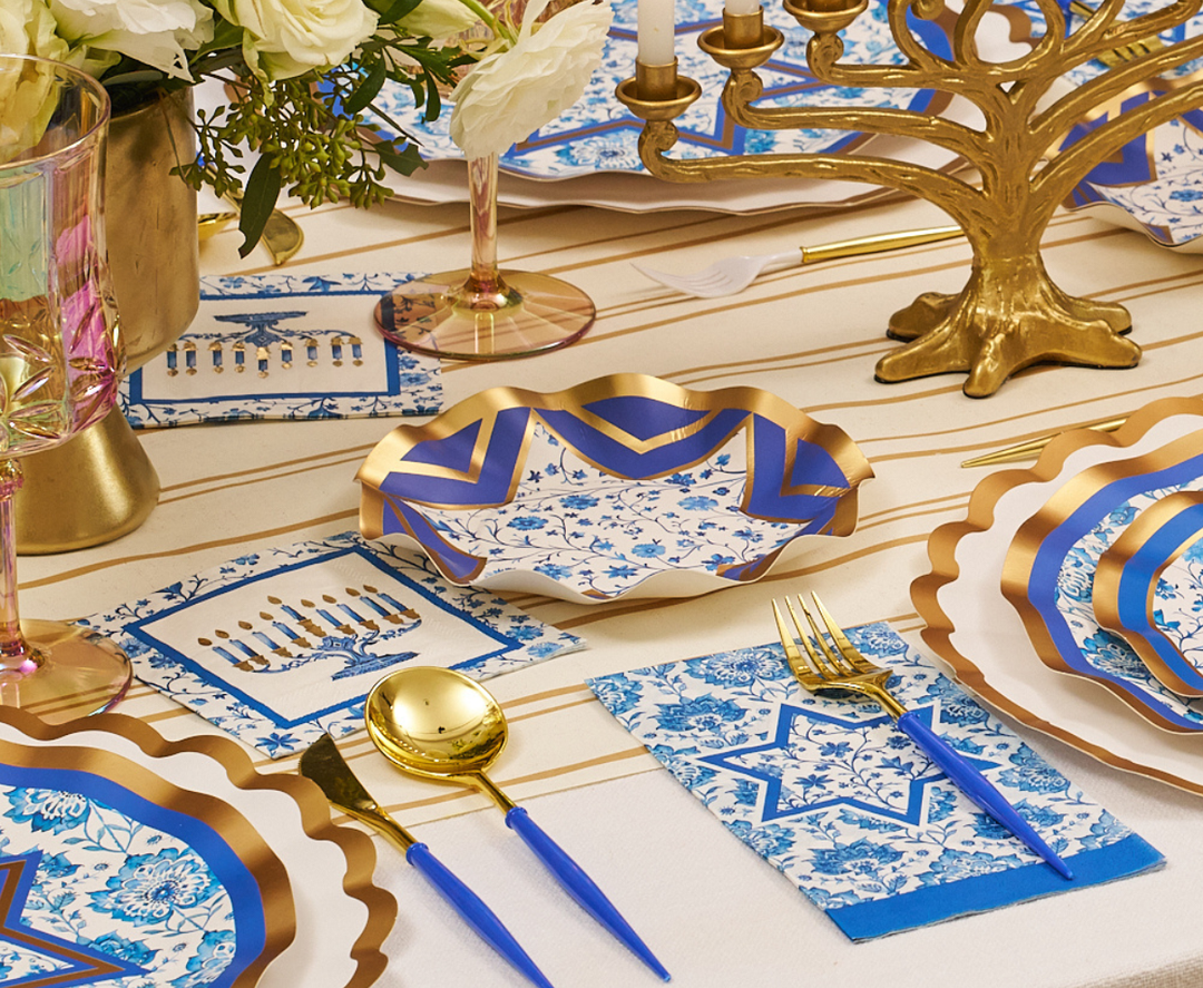 Create an elegant Hanukkah table setting with the Sophistiplate Hanukkah Toile Table Setting, featuring festive tablescape elements such as blue and gold designer paper plates, floral napkins, golden cutlery, menorah napkins, pink goblets, and a gold menorah centerpiece arranged on a striped tablecloth with white flowers.