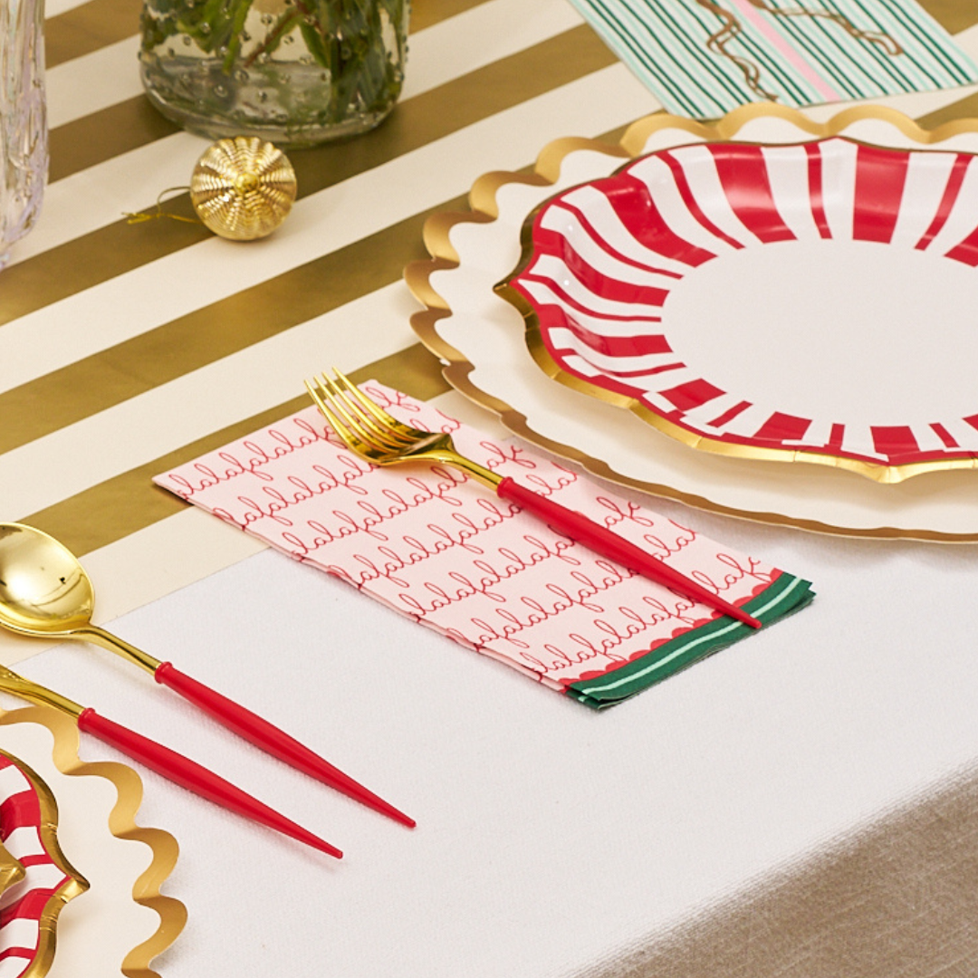 A festive table setting with gold and red utensils, scalloped edge plates featuring red stripes, and a gold-striped table runner atop a white tablecloth is beautifully accented by Sophistiplate’s Tis the Season Guest Towels (20pk), adding an extra touch of seasonal charm.