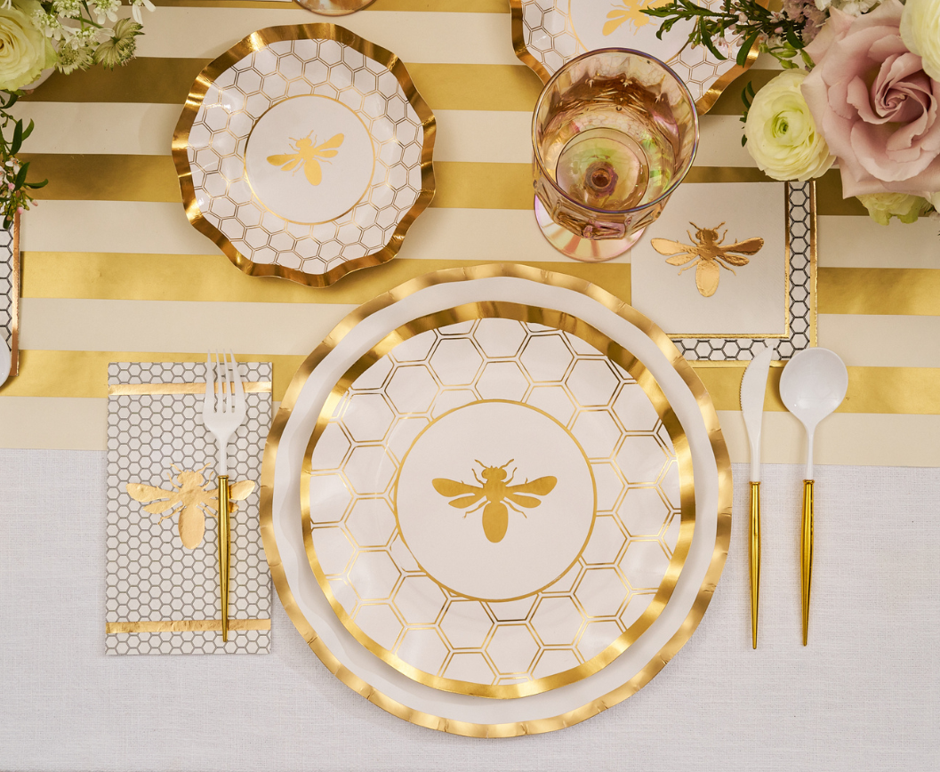 The White Honeybee Table Setting by Sophistiplate features elegant paper plates and napkins with bee and honeycomb designs, complemented by gold utensils, a striped table runner, and fresh flowers.