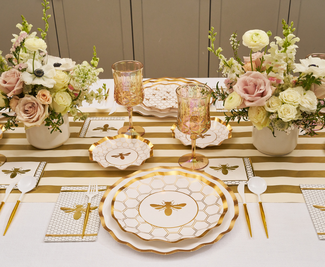 Create an elegant table setting with the Sophistiplate White Honeybee Table Setting, featuring gold and white bee-themed disposable plates, coordinating gold cutlery, pink glassware, and floral centerpieces of roses and greenery atop a striped table runner.