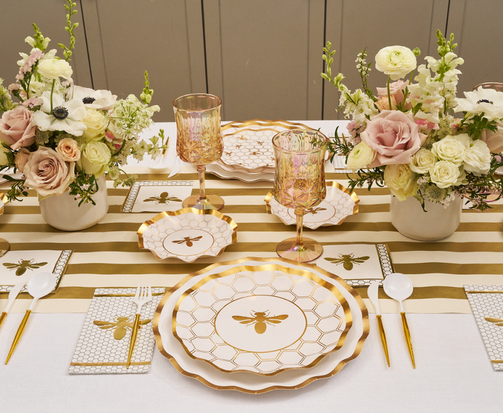 Create an elegant table setting with the Sophistiplate White Honeybee Table Setting, featuring gold and white bee-themed disposable plates, coordinating gold cutlery, pink glassware, and floral centerpieces of roses and greenery atop a striped table runner.