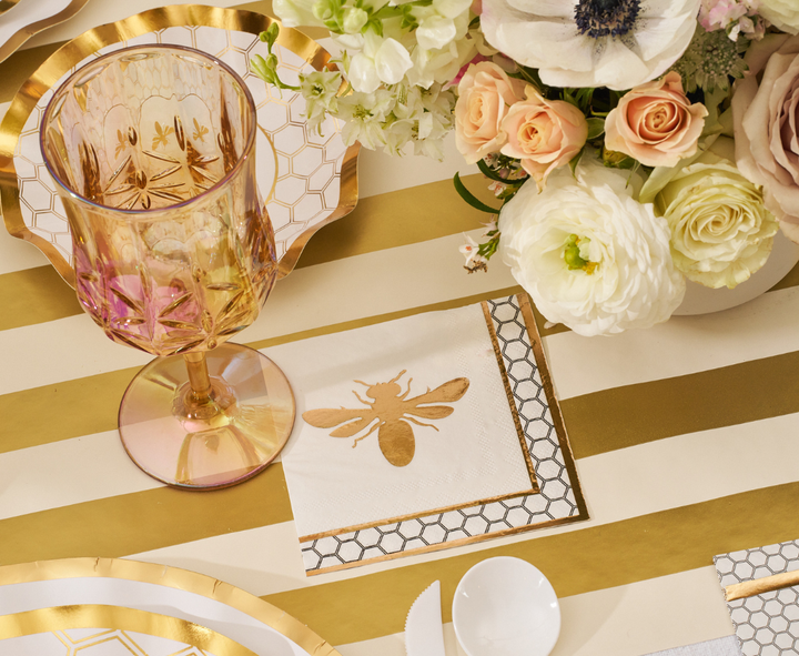 The White Honeybee Table Setting by Sophistiplate features pink-tinted goblets, bee-patterned napkins, floral arrangements, and elegant gold rim paper plates arranged on a white and gold striped tablecloth.
