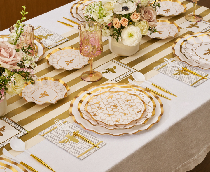 Create a refined table setting with the Sophistiplate White Honeybee Table Setting, featuring gold and white party plates, gold utensils, pink glasses, floral centerpieces, and bee-themed decorations atop a striped gold and white table runner.