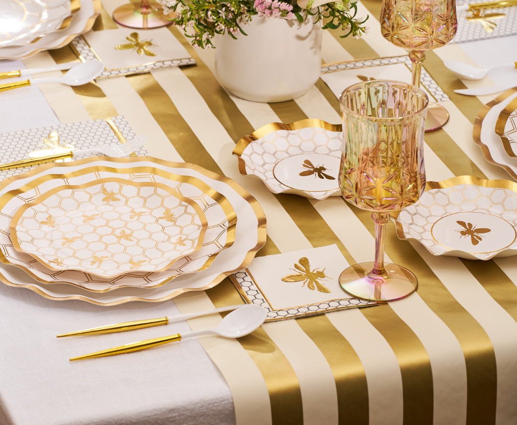 Transform your dining experience with the White Honeybee Table Setting from Sophistiplate, featuring a gold-striped tablecloth, bee-themed napkins, and elegant paper plates that highlight the sophisticated décor. Gold cutlery and disposable plates are paired with plastic spoons and pink-tinted glassware to create a charming atmosphere. A floral centerpiece completes the look, while a cohesive white and gold color scheme ties everything together.