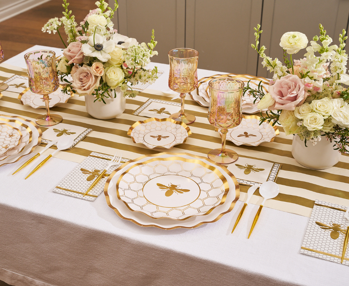 The Sophistiplate White Honeybee Table Setting features elegant paper plates with a bee motif, a gold-striped runner, gold utensils, blush and white floral centerpieces, and pink-tinted glasses, all arranged for a sophisticated formal meal.