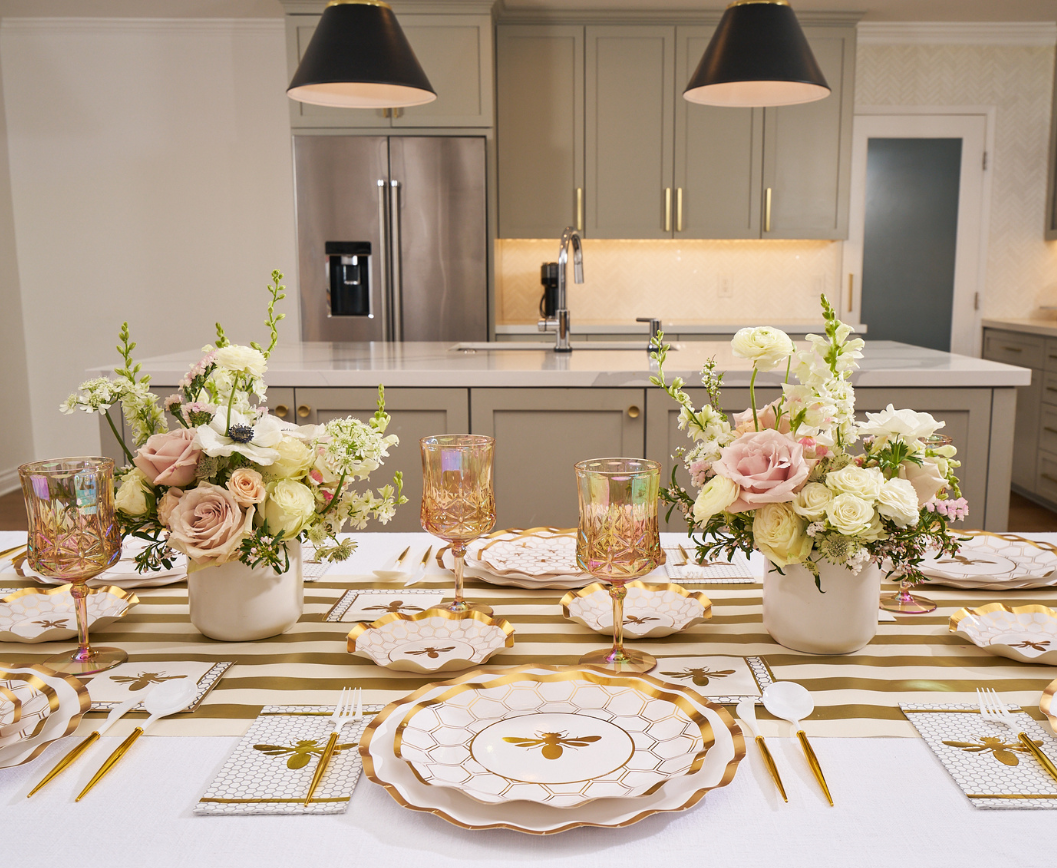 The Sophistiplate White Honeybee Table Setting, featuring elegant gold and white party plates, gold utensils, pink glasses, and floral centerpieces, is beautifully arranged in a modern kitchen with light gray cabinets and pendant lights.