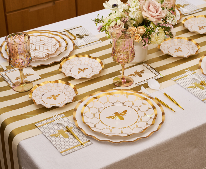 A White Honeybee Table Setting by Sophistiplate is arranged with elegant gold and white bee-themed disposable plates, honeycomb-patterned napkins that coordinate with the plates, a striped table runner, pink goblets, a floral centerpiece, and gold cutlery.