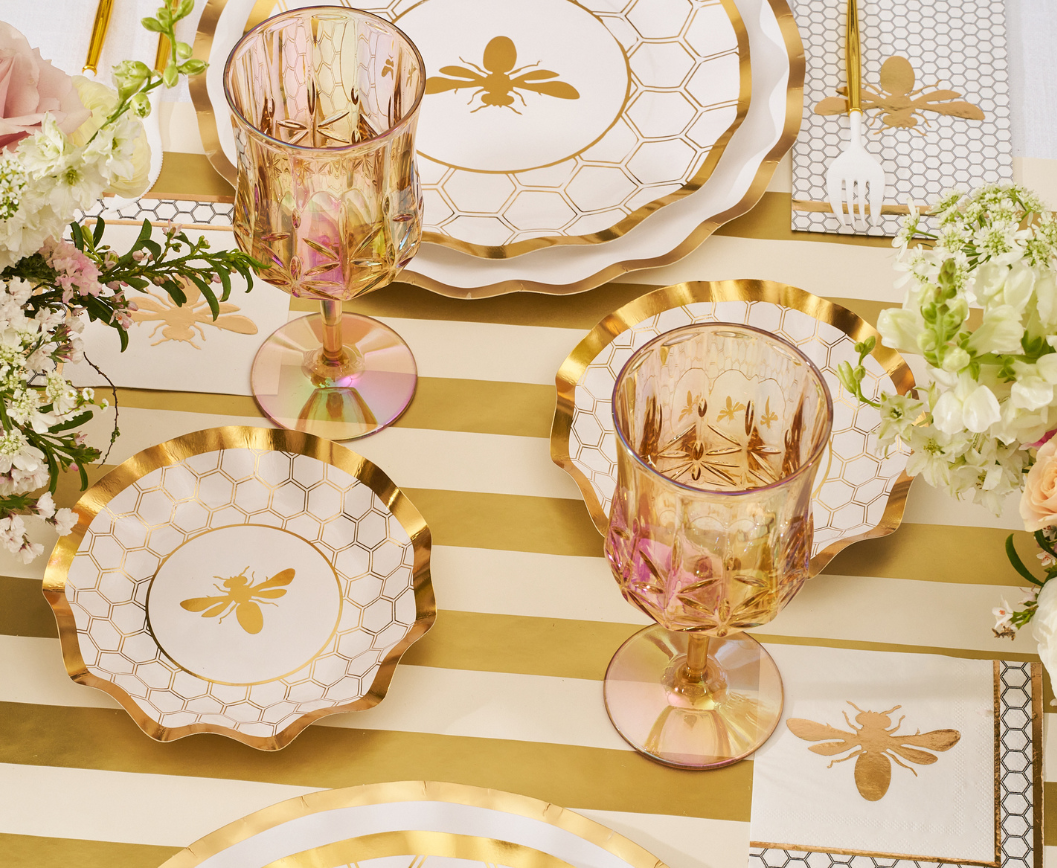The White Honeybee Table Setting by Sophistiplate features elegant gold and white party plates adorned with charming bee motifs, perfectly paired with gold-rimmed glasses, a striped tablecloth, floral centerpieces, and matching napkins embellished with bee designs. These stylish paper plates offer both sophistication and convenience for any special occasion.