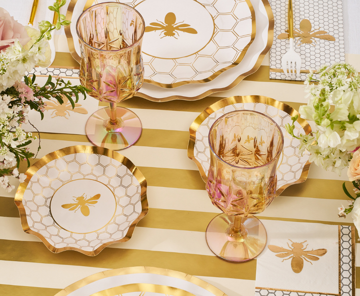 The White Honeybee Table Setting by Sophistiplate features elegant gold and white party plates adorned with charming bee motifs, perfectly paired with gold-rimmed glasses, a striped tablecloth, floral centerpieces, and matching napkins embellished with bee designs. These stylish paper plates offer both sophistication and convenience for any special occasion.