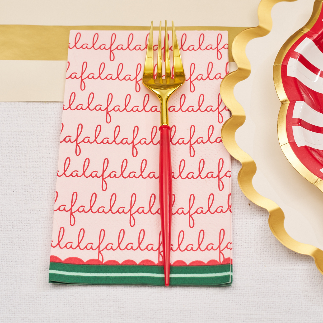 A gold and red fork rests on a festive “Tis the Season” guest towel from Sophistiplate, featuring “falalala” repeated in red script, placed next to a red and white striped plate with a gold scalloped edge on a decorated table.