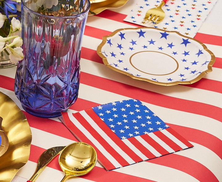 A festive table is set with an American flag napkin, gold utensils, a blue glass, star plates, and Sophistiplate’s Party in the USA Appetizer/Dessert Bowls/8pk atop a red-and-white striped tablecloth for a patriotic look.