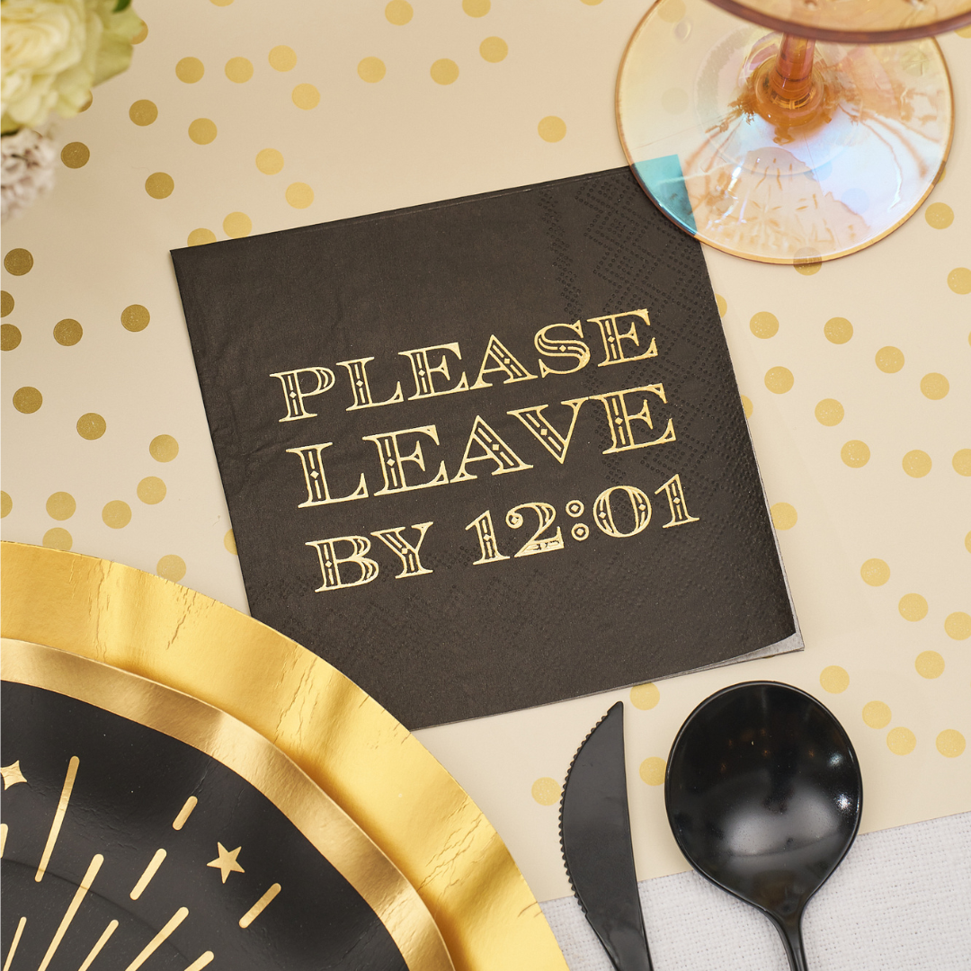 A black Midnight Sparkle Paper Cocktail Napkin Please Leave By 12:01 /26pk with gold-printed text, arranged among gold-rimmed plates and cutlery.