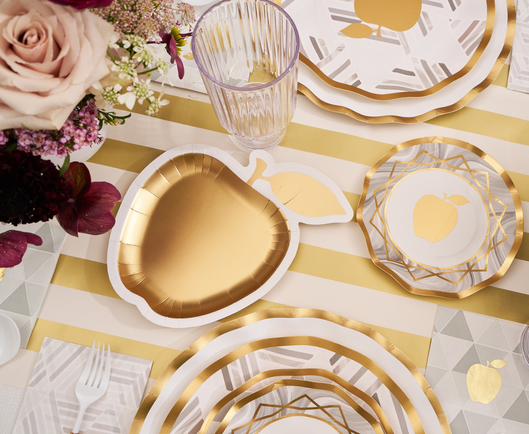 The Sophistiplate Golden Apple Table Setting features a gold apple-shaped plate, a beautiful floral arrangement, a striped tablecloth, and coordinated disposable plates, cups, and cutlery—offering an elegant and festive option for celebrating Rosh Hashanah.