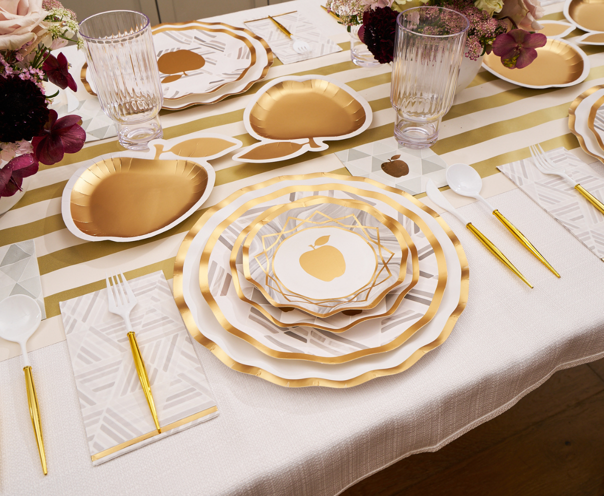 Sophistiplate’s Golden Apple Table Setting disposable plates in gold and white, adorned with gold apple designs, are paired with coordinating gold utensils, clear glasses, and floral centerpieces on a striped table runner—creating an elegant setting perfect for celebrating Rosh Hashanah in style.
