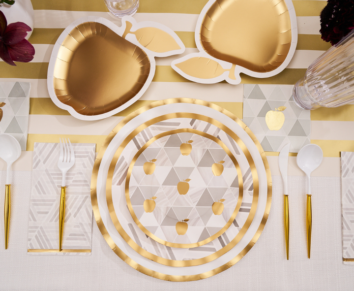The Sophistiplate Golden Apple Table Setting is ideal for Rosh Hashanah, showcasing gold apple-shaped disposable plates, a round plate adorned with gold apple designs, elegant gold cutlery, and geometric napkins—all beautifully displayed on a white and gold striped tablecloth.