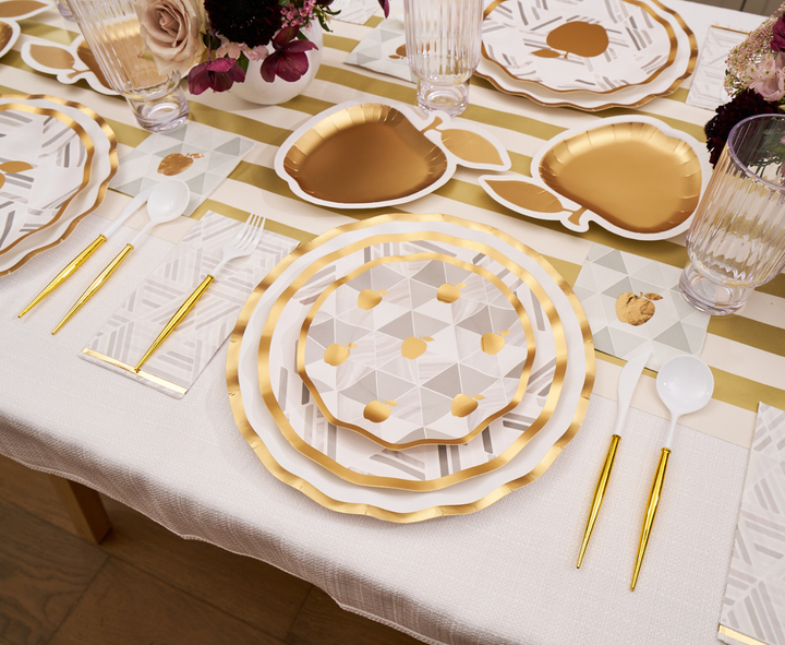 Celebrate Rosh Hashanah in style with the Sophistiplate Golden Apple Table Setting, which includes gold-rimmed geometric plates, elegant disposable plates, gold heart-shaped plates, gold flatware, clear ribbed glasses, and a striped white and gold table runner adorned with floral centerpieces for a festive touch.