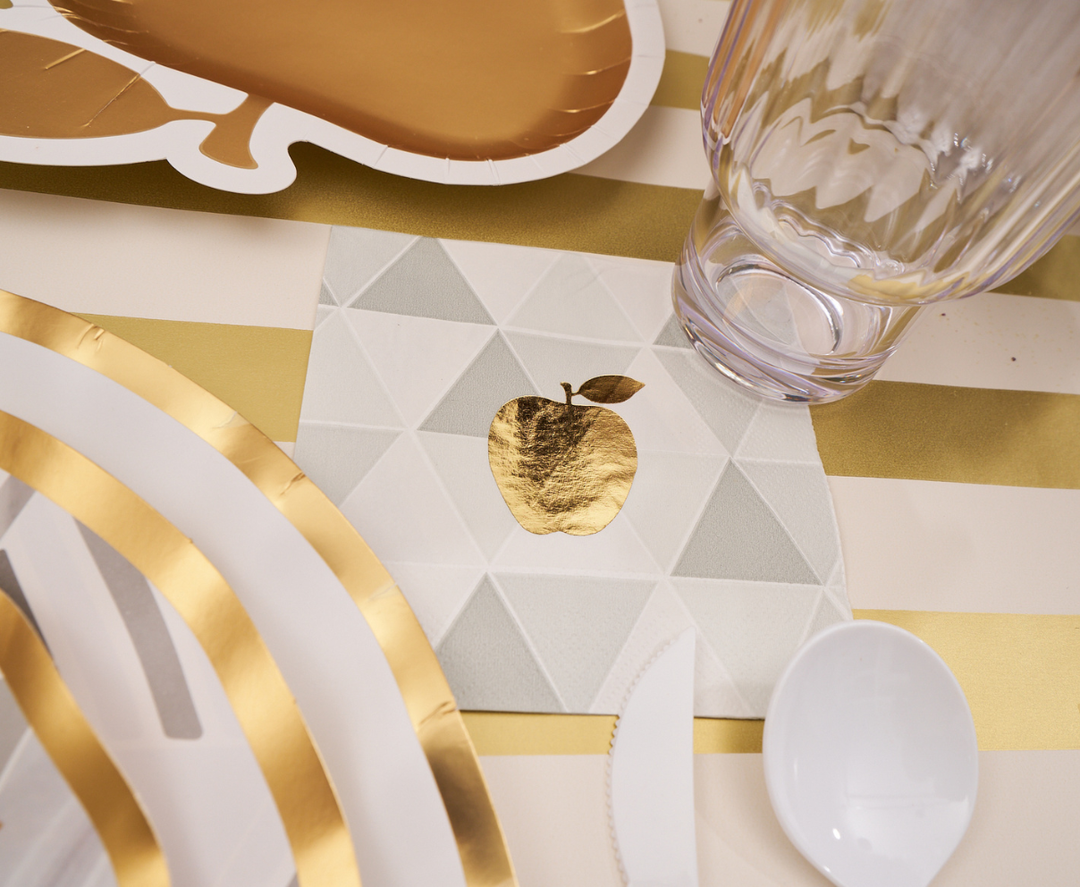 Elevate your Rosh Hashanah celebration with the Sophistiplate Golden Apple Table Setting, featuring a gold-edged disposable plate, utensils, clear glass, and a napkin adorned with a shiny gold apple on a geometric background—all in an elegant white and gold striped design.
