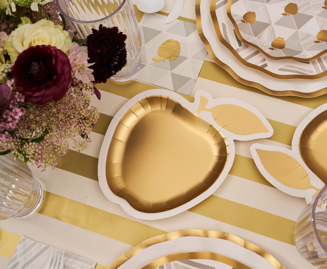 A gold, apple-shaped disposable plate from Sophistiplate’s Golden Apple Table Setting collection rests on a striped gold and white tablecloth, surrounded by floral arrangements, clear cups, and other patterned plates at a festive Rosh Hashanah table.