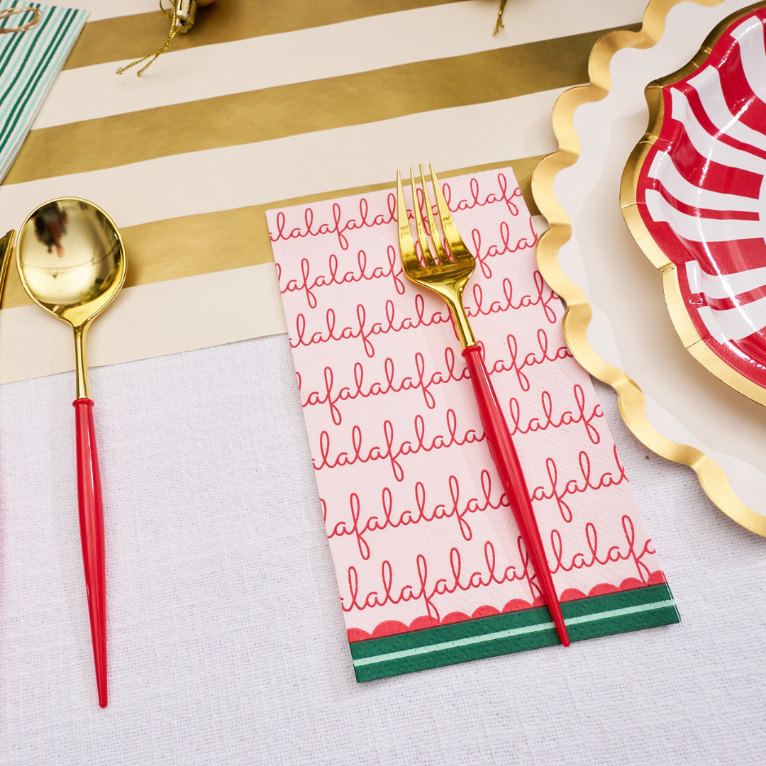 A festive table setting with a gold spoon and fork, a red and white striped plate, a scalloped plate with gold trim, and a Sophistiplate Tis the Season Guest Towel napkin featuring bold red “falalala” text and green borders, all set atop a white and gold striped table runner.