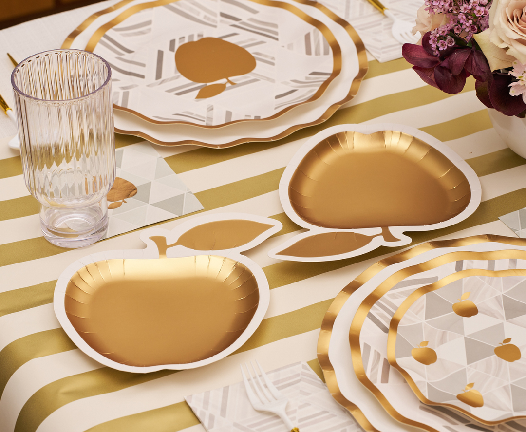 Introducing the Golden Apple Table Setting by Sophistiplate—ideal for Rosh Hashanah celebrations. This elegant set features a gold and white striped tablecloth, golden apple-shaped disposable plates, geometric and polka-dot accent plates, ribbed glassware, gold utensils, and a beautiful floral centerpiece to complete your festive table.