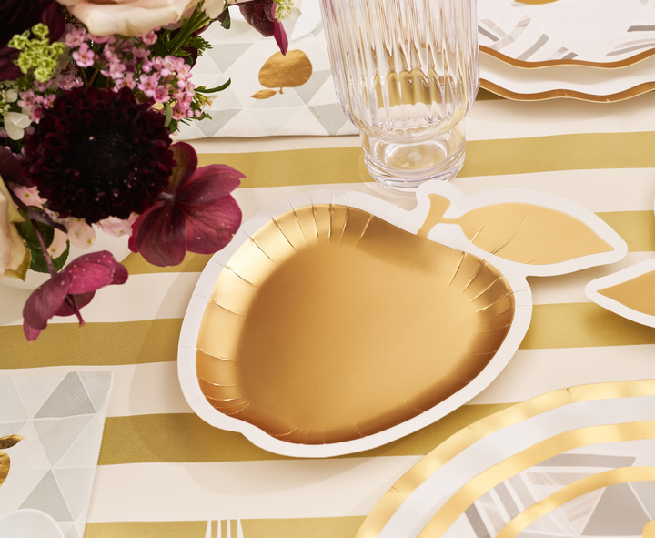 The Sophistiplate Golden Apple Table Setting includes a gold apple-shaped disposable plate displayed on a striped tablecloth, accented by glassware, flowers, and coordinating party tableware—ideal for adding festive style to occasions such as Rosh Hashanah.