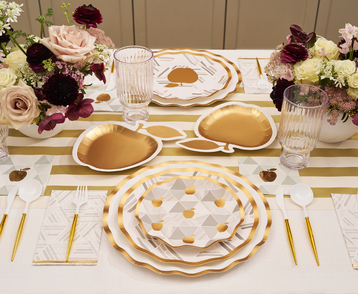 A Sophistiplate Golden Apple Table Setting showcases an elegant Rosh Hashanah celebration table with gold and white disposable plates, gold cutlery, paper napkins, clear glasses, and floral centerpieces featuring pink and burgundy flowers atop a gold-striped table runner.