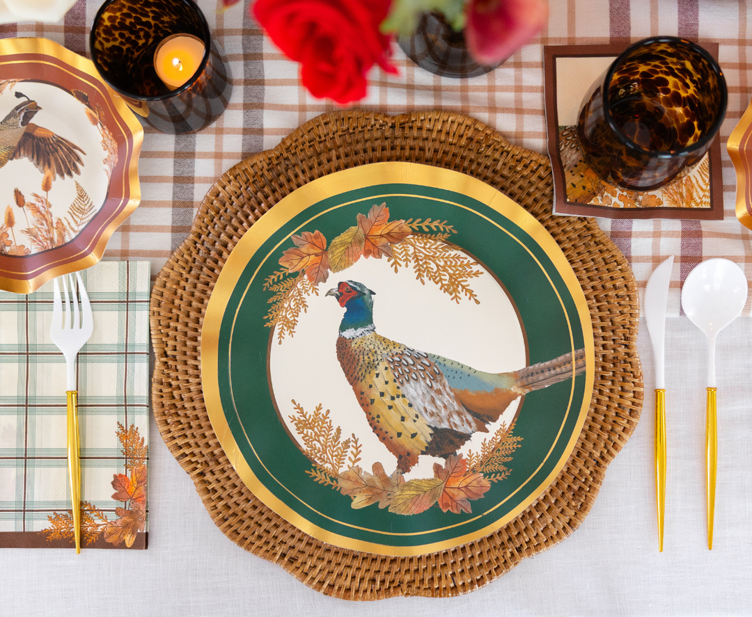 Create a festive ambiance with the Sophistiplate Woodland Harvest Table Setting—a designer ensemble featuring pheasant-themed paper plates, wicker placemats, gold utensils, plaid napkins, tortoiseshell glasses, candles, and red floral accents—all beautifully arranged on a plaid tablecloth.