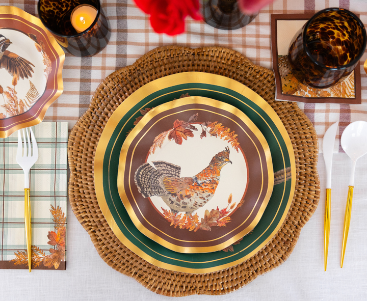 Table setting featuring Sophistiplate Woodland Harvest Salad Plates (8-pack) adorned with pheasant and autumn leaf motifs, arranged on a woven placemat with gold-accented flatware, plaid napkin, amber glassware, a candle, and a plaid fall-themed tablecloth.