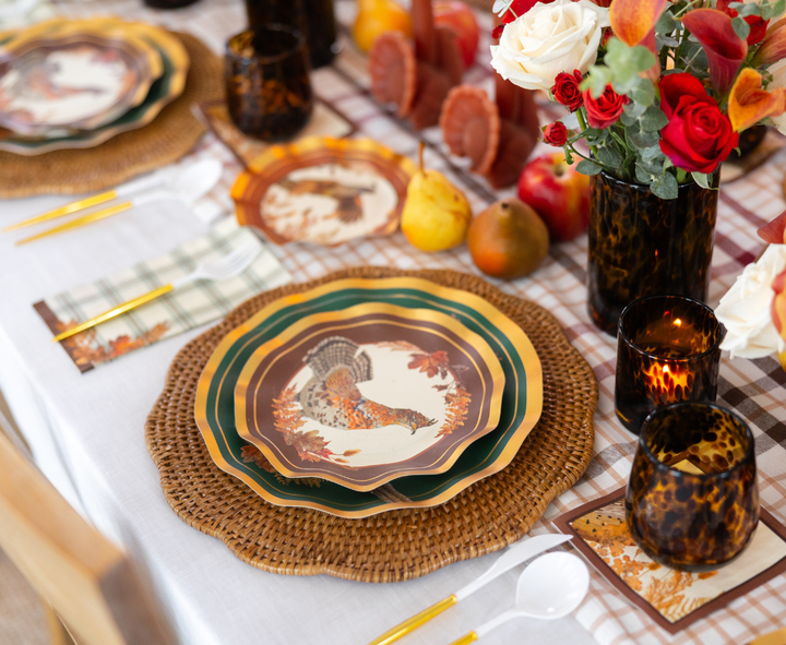 The Woodland Harvest Table Setting by Sophistiplate creates a festive harvest tablescape with layered turkey-themed designer paper plates, gold utensils, plaid napkins, brown glasses, a centerpiece of red and white flowers, decorative pears, and a checkered tablecloth.
